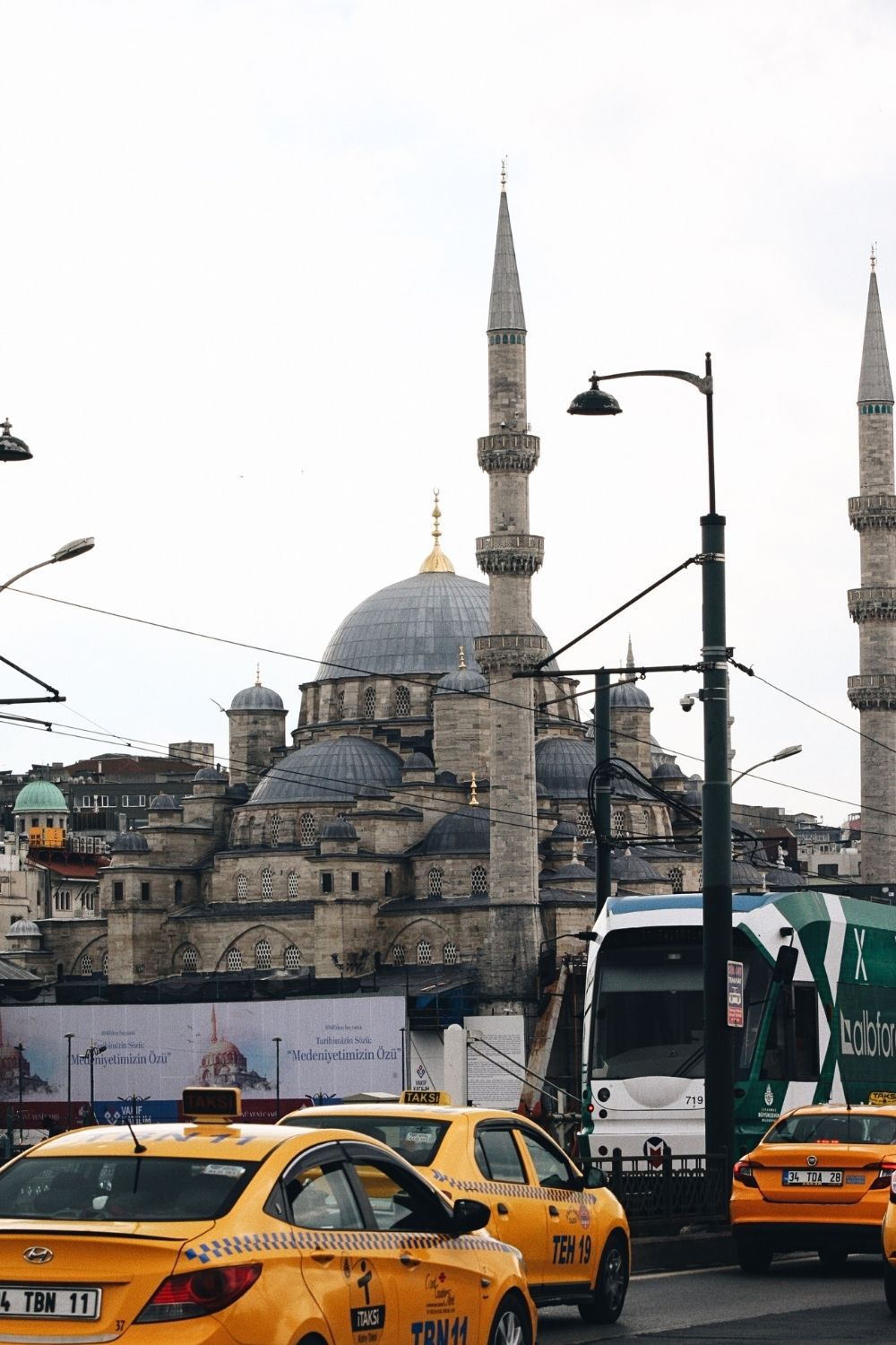 This image features a bustling street scene in Istanbul, with the iconic domes and minarets of a historic mosque, possibly the Yeni Cami (New Mosque), in the background. Yellow taxis and a green tram dominate the foreground, illustrating the mix of modern transportation against a backdrop of Ottoman-era architecture