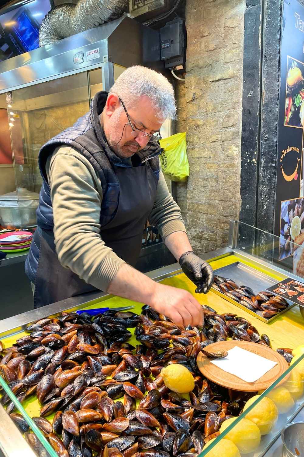 The image features a vendor preparing freshly stuffed mussels, or "midye dolma," a popular Istanbul street food, at a well-lit stall. The mussels are displayed alongside bright yellow lemons, ready to be squeezed.