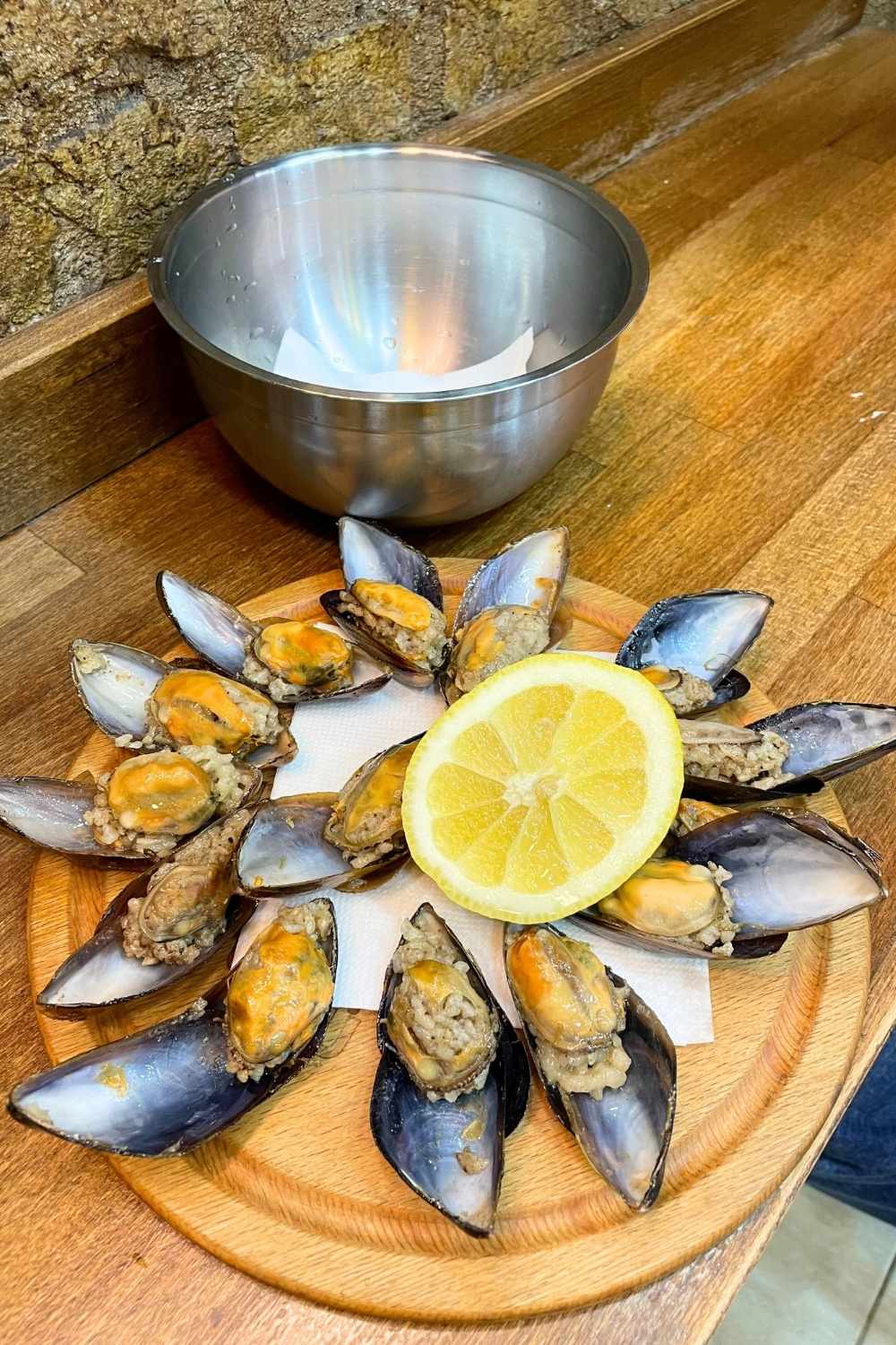 The image showcases a wooden plate of freshly prepared stuffed mussels, or "midye dolma," arranged in a circular pattern with a slice of lemon in the center for squeezing.