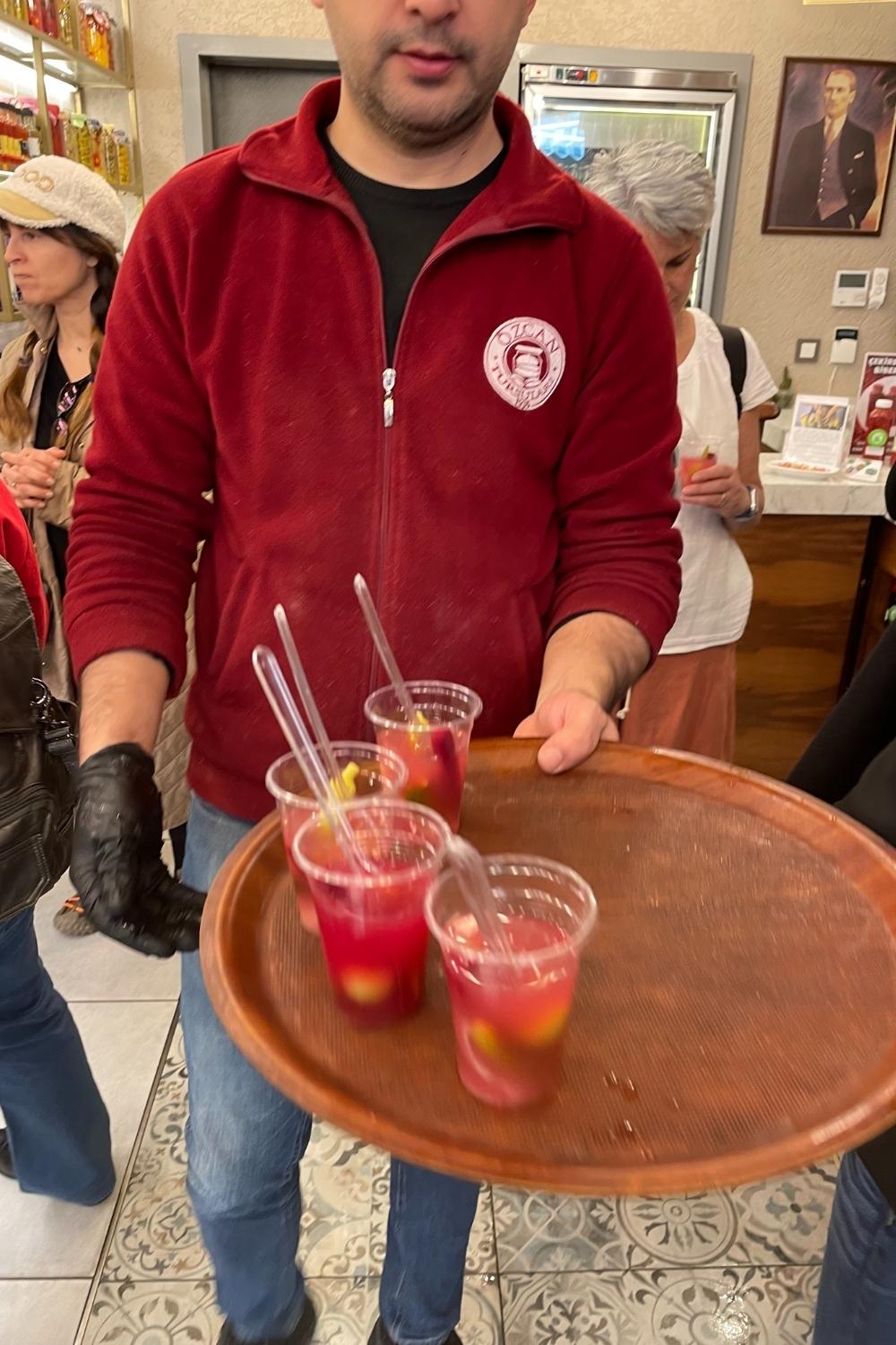 The image features a server holding a tray with cups of vibrant red pickle juice, garnished with whole pickles and vegetables, ready to be served.