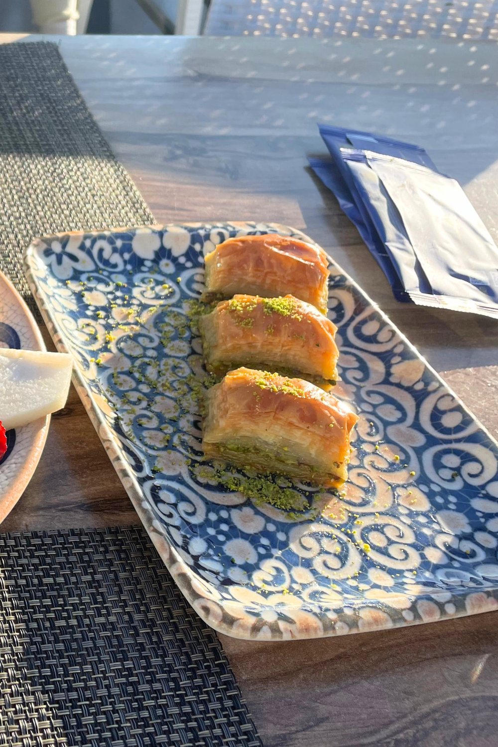 The image features a beautifully arranged plate of Turkish baklava on a decorative blue-patterned dish. The golden, flaky layers of pastry are topped with a sprinkle of ground pistachios, highlighting this iconic dessert from Istanbul's rich culinary tradition.