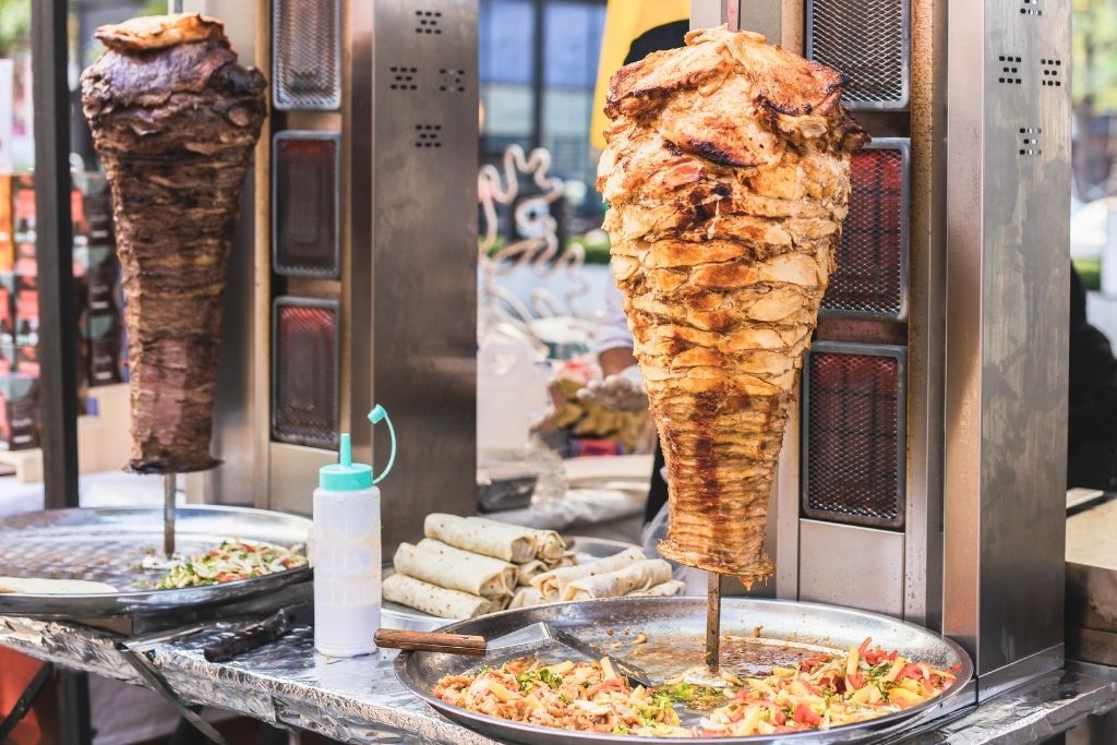 The image features two rotating spits of döner kebab, one with juicy grilled beef and the other with golden chicken, surrounded by a variety of fresh toppings and wraps.