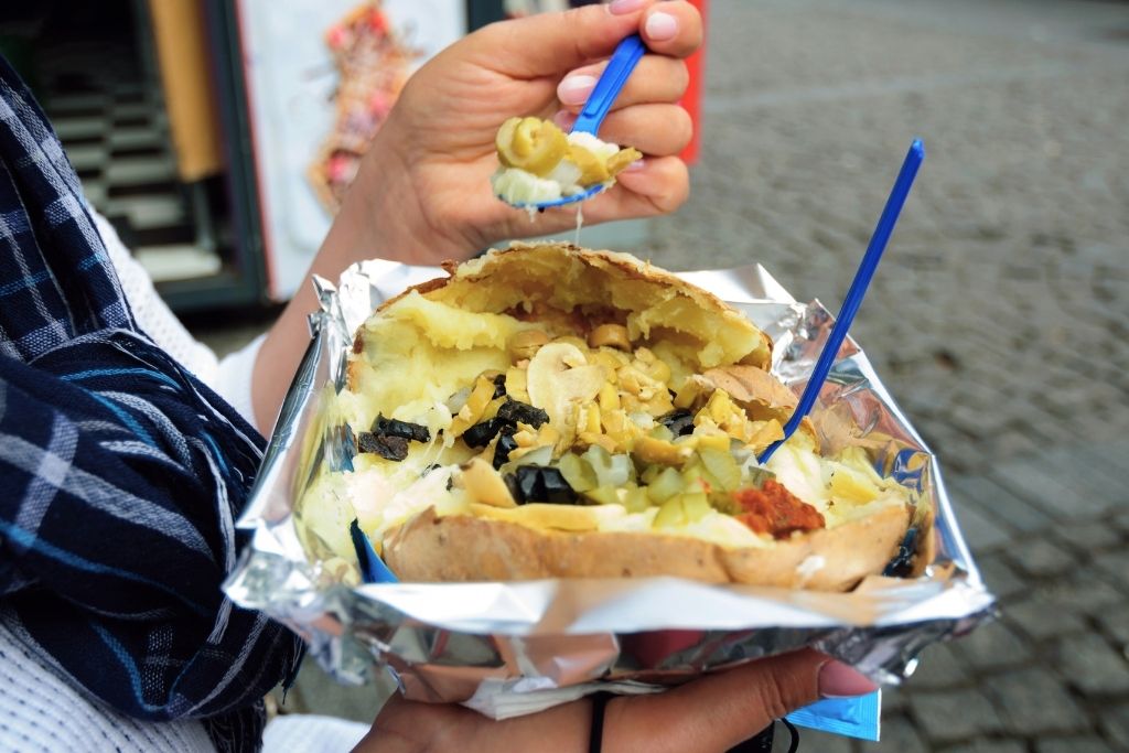 The image showcases a serving of kumpir, a loaded baked potato, stuffed with a variety of toppings such as olives, pickles, corn, mushrooms, and creamy sauces. Wrapped in foil, this popular Turkish street food offers a hearty meal.