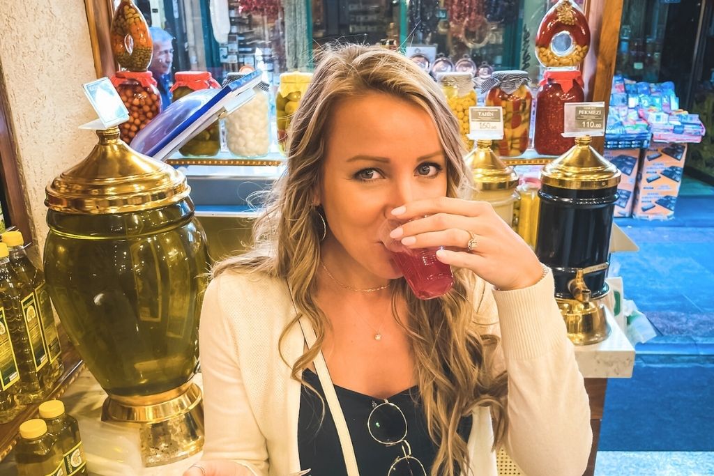 The image shows Kate enjoying a glass of vibrant pickle juice at a spice and condiment shop in Istanbul, surrounded by jars of colorful pickles, tahini, and molasses on display. The scene highlights a unique and authentic experience of Istanbul street food culture, perfect for adventurous travelers.