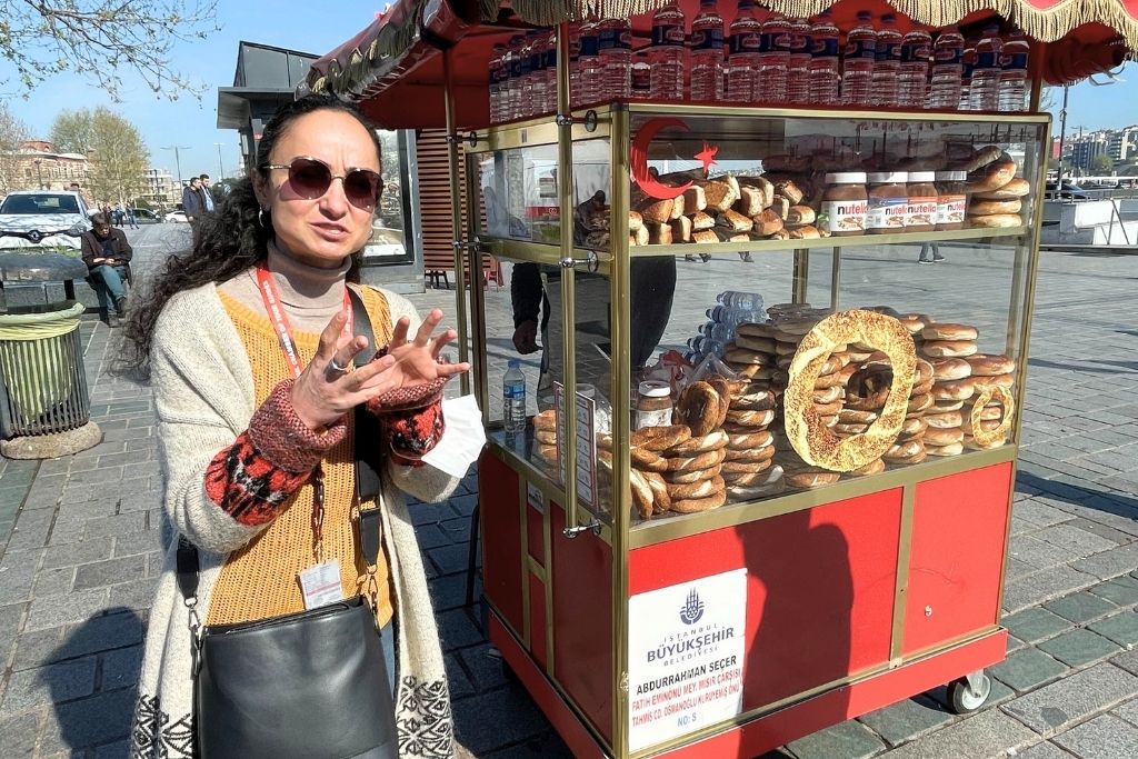 The image features a woman standing next to a red street vendor cart in Istanbul, selling freshly baked simit, a traditional Turkish sesame bagel-like bread. The cart is adorned with neatly stacked simit, water bottles, and jars of Nutella.