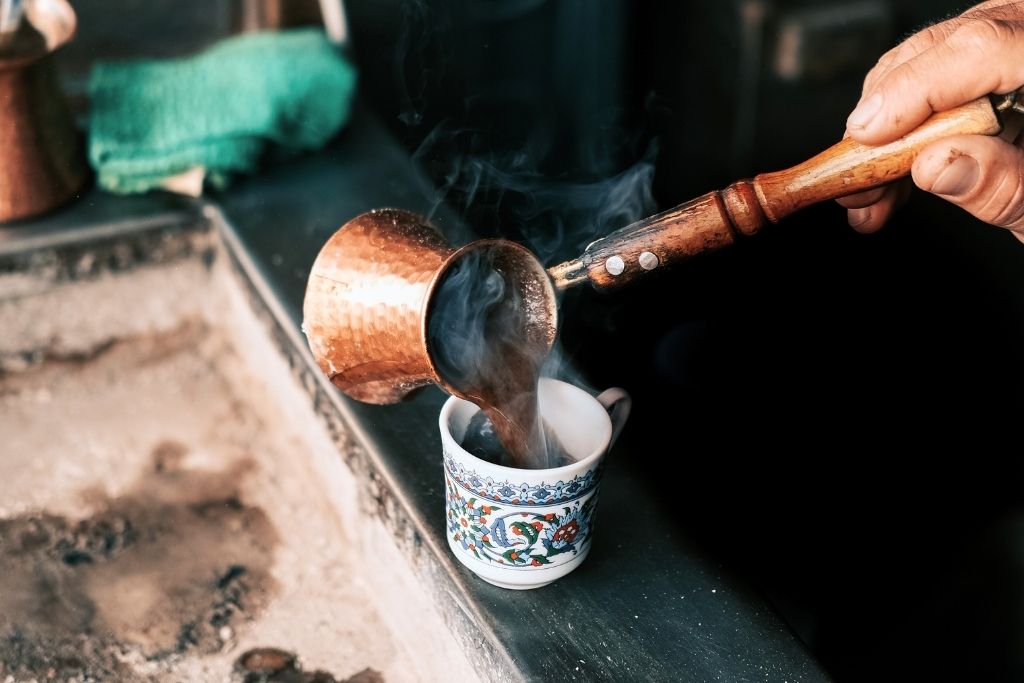 The image shows a steaming cup of Turkish coffee being poured from a traditional copper cezve into a decorative porcelain cup. The rich aroma and authentic preparation method capture the essence of Istanbul's deep-rooted coffee culture.