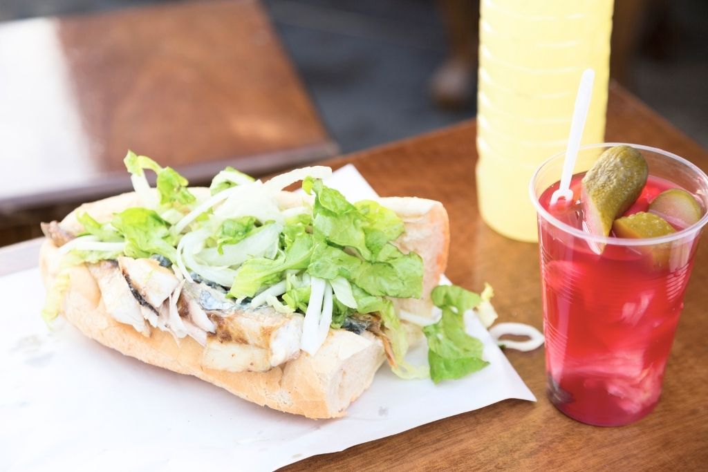 The image showcases a traditional Turkish balık-ekmek, a grilled fish sandwich served on a baguette with fresh lettuce and onions, paired with a vibrant cup of pickle juice featuring pickles and vegetables.