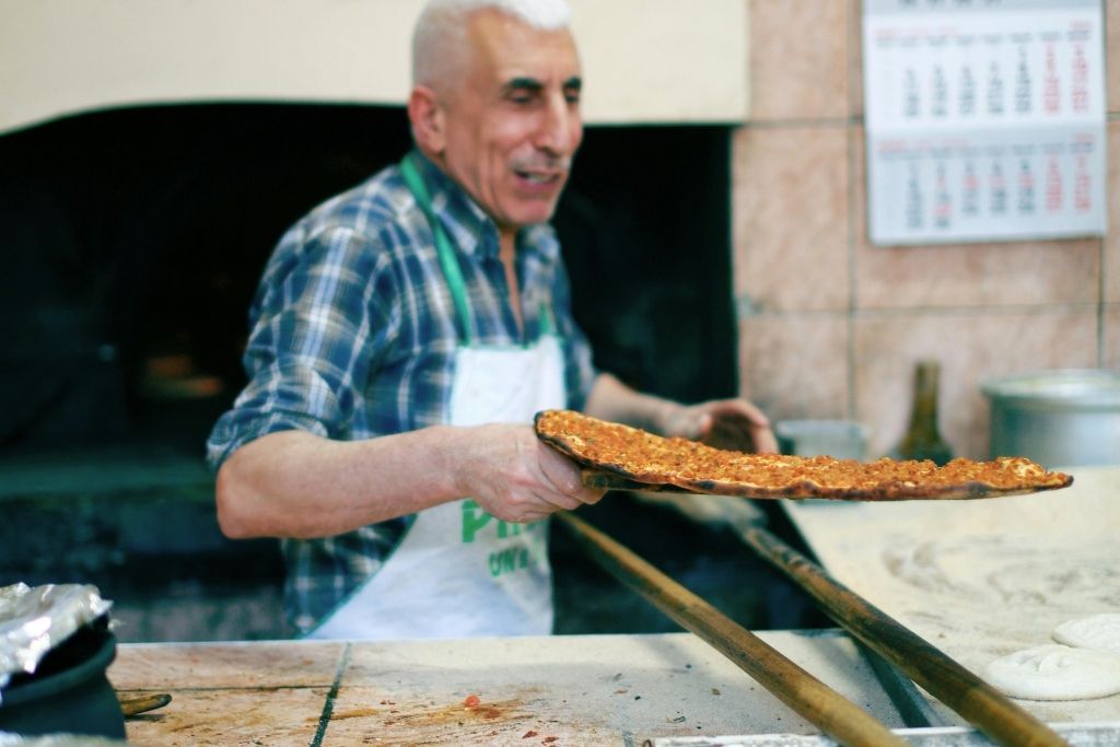 The image captures a smiling vendor serving freshly baked lahmacun, also known as Turkish street pizza, directly from a traditional wood-fired oven. The thin, crispy flatbread topped with minced meat, herbs, and spices exemplifies the authentic and flavorful street food culture of Istanbul.