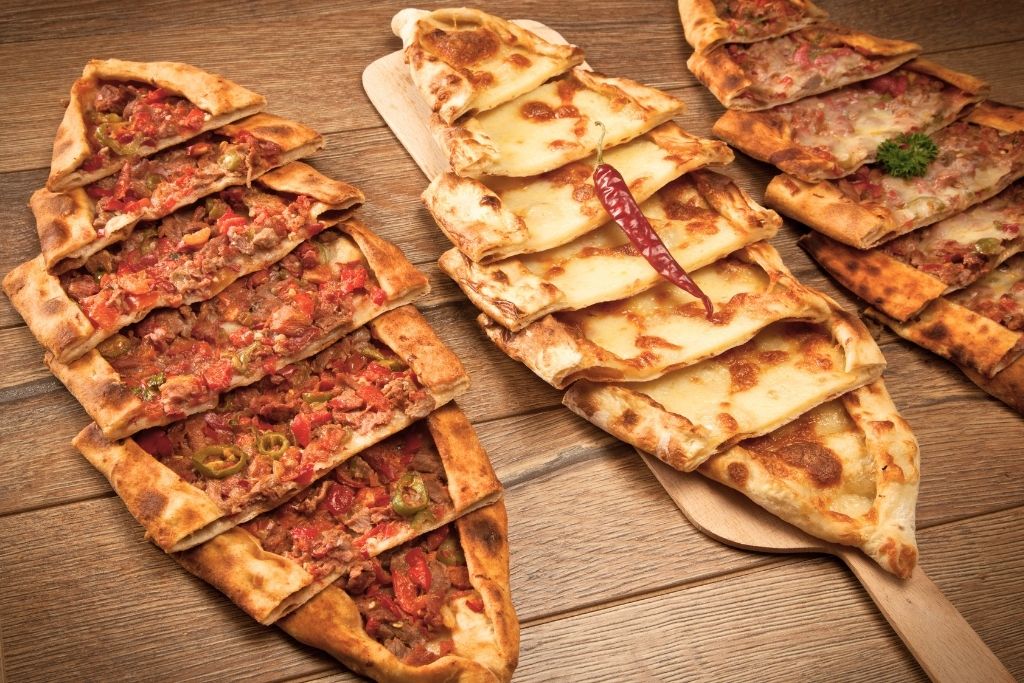 The image showcases traditional Turkish pide, often referred to as "Turkish pizza," served on a wooden surface. The dish includes various toppings like minced meat with peppers and tomatoes, melted cheese, and spiced meats, capturing the diversity and flavors of Istanbul's street food cuisine.