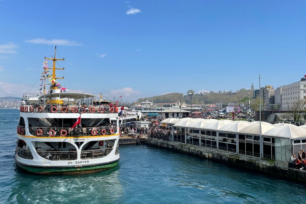 This image depicts a ferry terminal in Istanbul, with a large white ferry named "ŞH SARIYER" docked at the pier. The ferry is adorned with colorful flags, and the surrounding area is bustling with people under a bright blue sky. The waterfront scene captures the energy of daily life by the Bosphorus, with city buildings and greenery visible in the background.