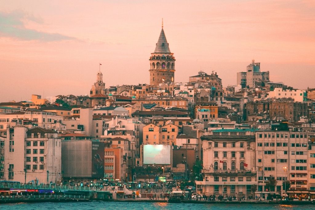 This stunning image captures a sunset view of Istanbul's cityscape, with the iconic Galata Tower standing tall amidst a cluster of historic and modern buildings. The warm hues of the sunset sky cast a soft, golden glow over the scene, highlighting the architectural charm and vibrant energy of the city. The waterfront in the foreground adds depth, with reflections shimmering in the water, completing the picturesque atmosphere.