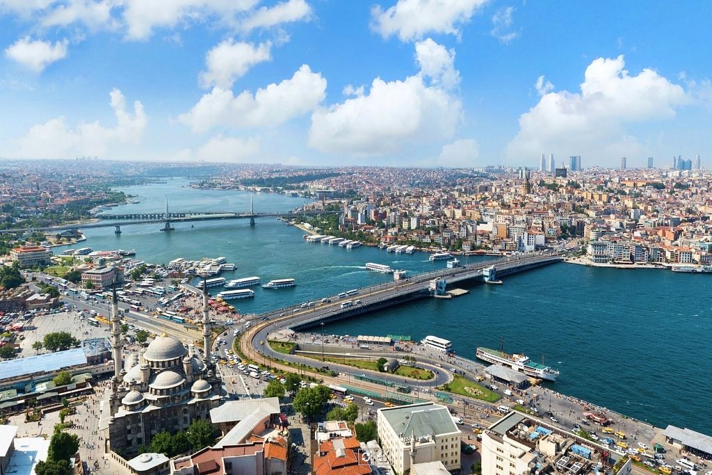 This image shows a vibrant aerial view of Istanbul, featuring the Golden Horn and Bosphorus waters framed by iconic bridges, such as the Galata Bridge. The foreground includes historic architecture, like mosques with domes and minarets, while the background displays a sprawling cityscape under a clear blue sky with fluffy white clouds.