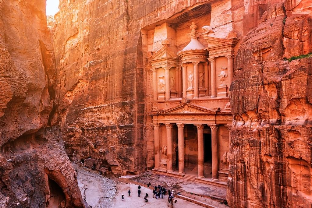 This photo showcases the iconic Al-Khazneh, also known as the Treasury, in Petra, Jordan. Carved into the reddish sandstone cliffs, the ancient Nabatean structure is illuminated by soft sunlight, emphasizing its intricate details and grandeur. Visitors at the base provide a sense of scale, highlighting the monument's impressive size.