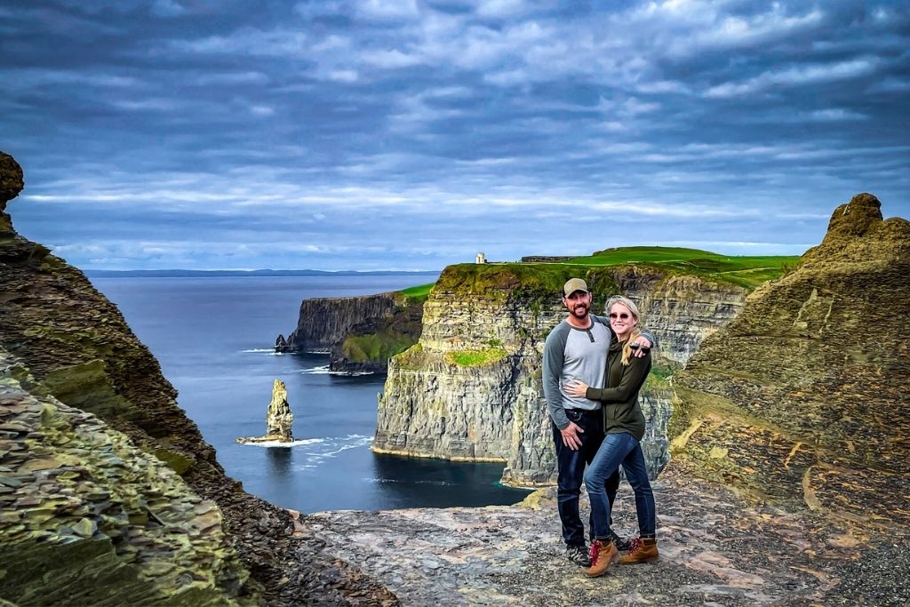 This photo features the breathtaking Cliffs of Moher in Ireland, showcasing dramatic rock formations towering above the Atlantic Ocean. Kate and her husband poses on a rugged cliff edge, surrounded by lush green grass and expansive ocean views under a cloudy sky.