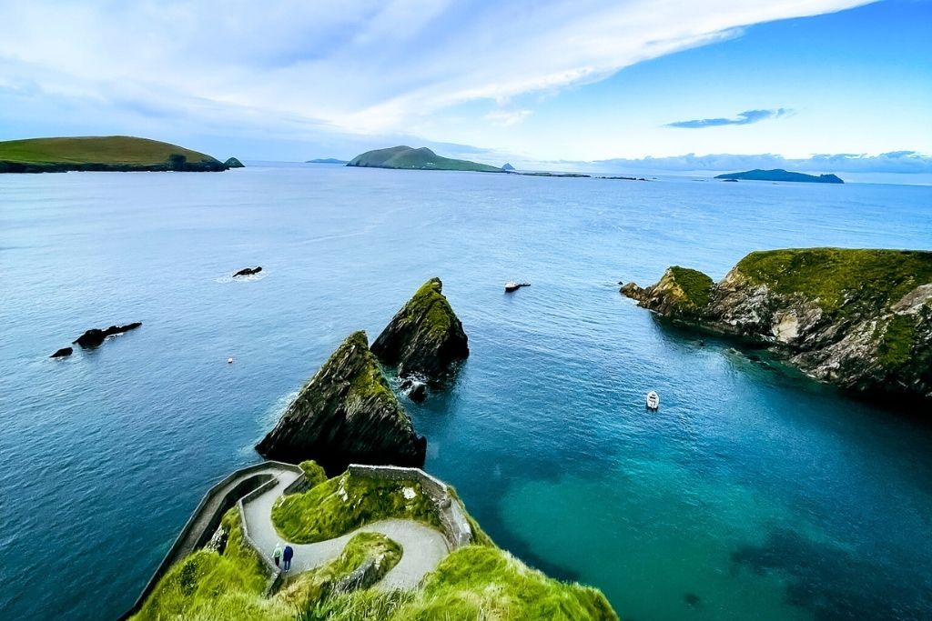 This image captures the stunning Dingle Peninsula in Ireland, featuring a winding pathway leading to rocky outcrops surrounded by vibrant blue waters. The scenic view showcases lush green cliffs and islands in the distance under a partly cloudy sky.