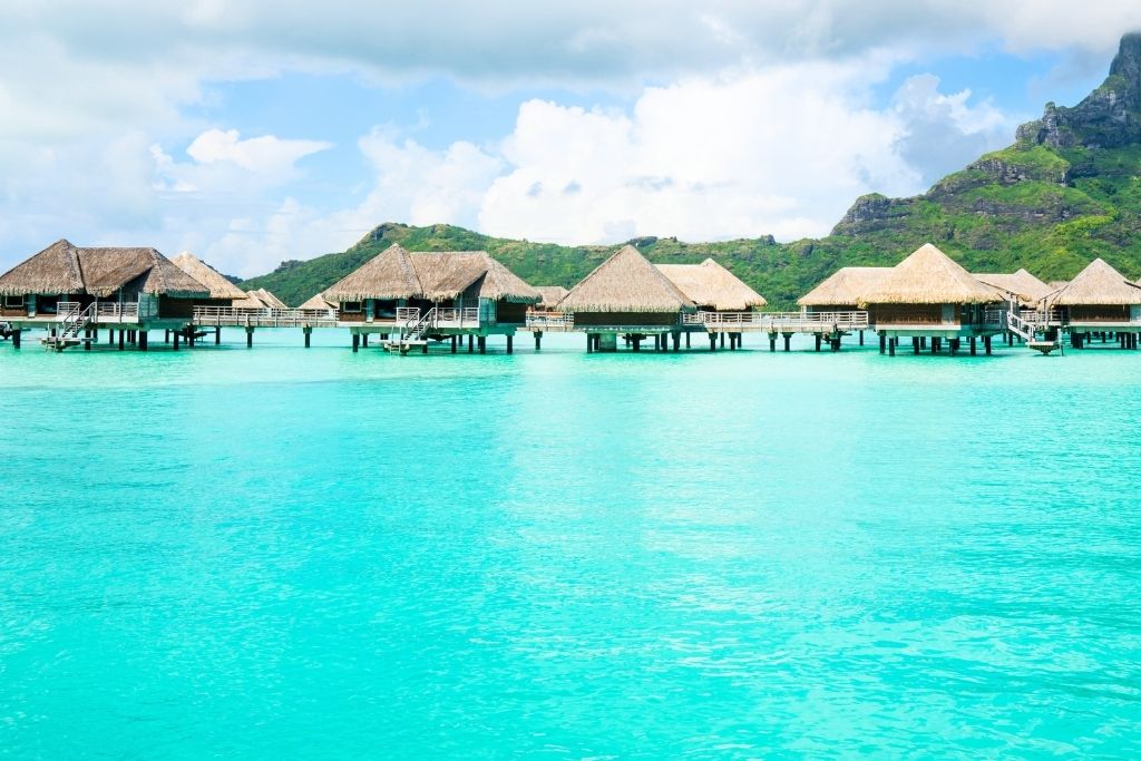 This image features overwater bungalows in Moorea, French Polynesia, set against a backdrop of lush green mountains and a vibrant turquoise lagoon. The thatched-roof villas stand on stilts above the crystal-clear water, offering a serene and idyllic tropical retreat. Soft white clouds and the natural scenery create a postcard-perfect view of paradise.