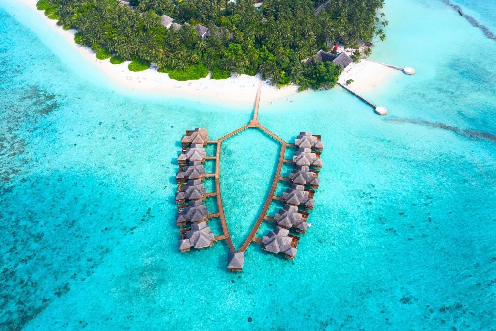 This aerial photo showcases luxurious overwater villas arranged in a symmetrical pattern on a turquoise lagoon, connected by wooden walkways. The backdrop features a lush green island with white sandy beaches, perfectly embodying the serene beauty of the Maldives.