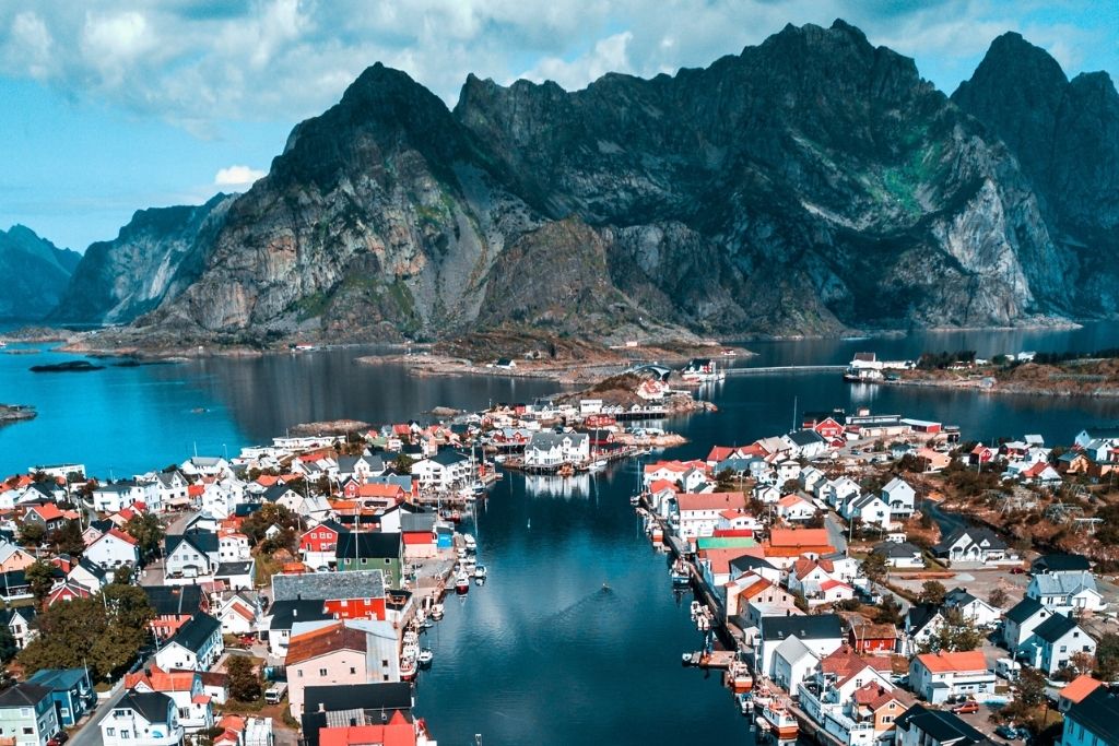 This image showcases a picturesque Norwegian fishing village nestled between a calm, reflective fjord and towering, rugged mountains. The colorful houses and boats along the waterfront create a vibrant contrast against the natural beauty of the surrounding landscape.