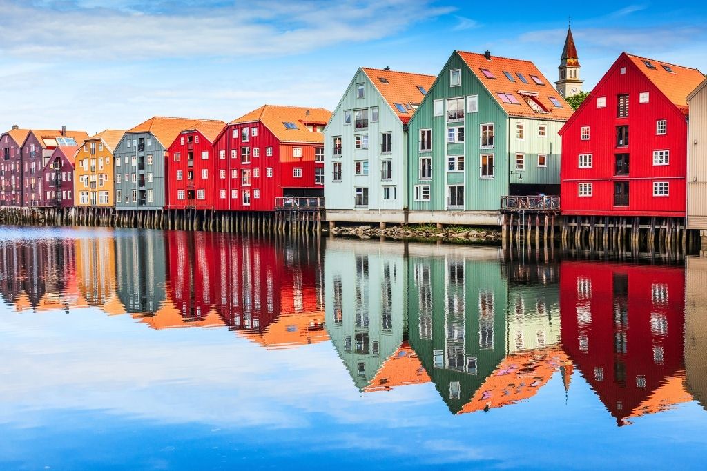 This image features colorful wooden houses lining the waterfront in Norway, their vibrant facades reflecting perfectly on the calm, glass-like water below. The combination of bright red, pastel green, and mustard tones against a blue sky creates a lively yet serene atmosphere, showcasing the charm of Scandinavian architecture.