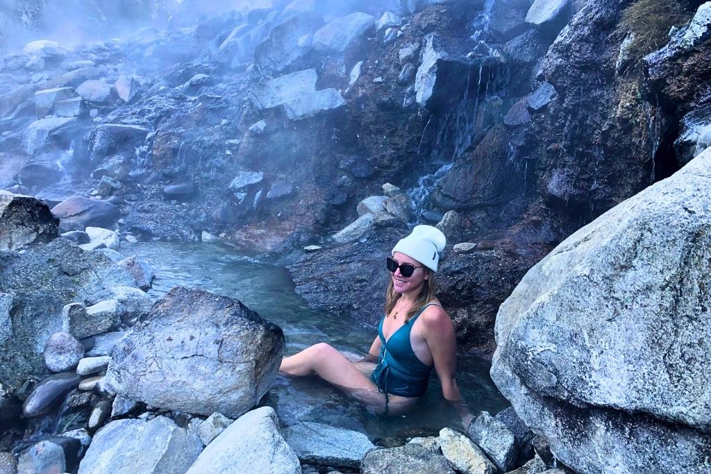 Kate in a green swimsuit, white beanie, and sunglasses relaxes in a natural hot spring surrounded by large rocks and cascading steamy water in Idaho.