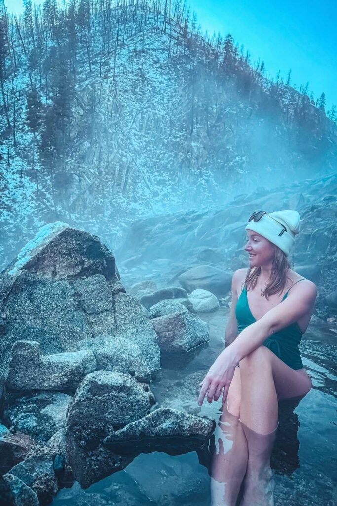 in a green swimsuit and white beanie sits in the steaming Pine Flats Hot Springs, surrounded by rugged rocks and a misty, snow-covered mountain landscape.