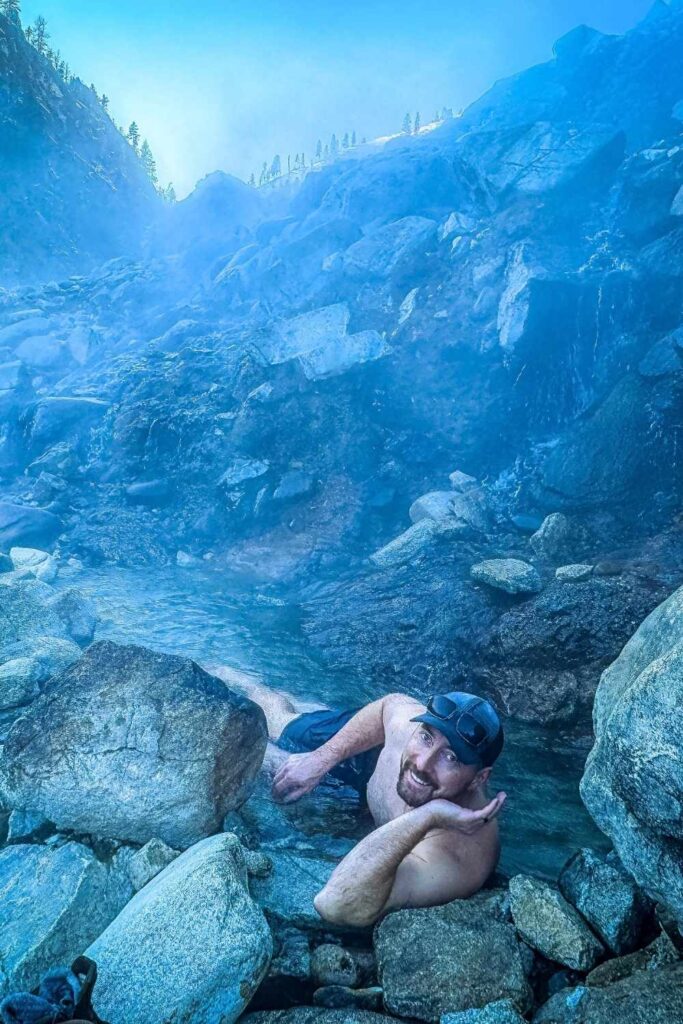 Kates husband in sunglasses and swim trunks lounges in the warm waters of Pine Flats Hot Springs, surrounded by misty, rocky terrain with steam rising from the geothermal pools.