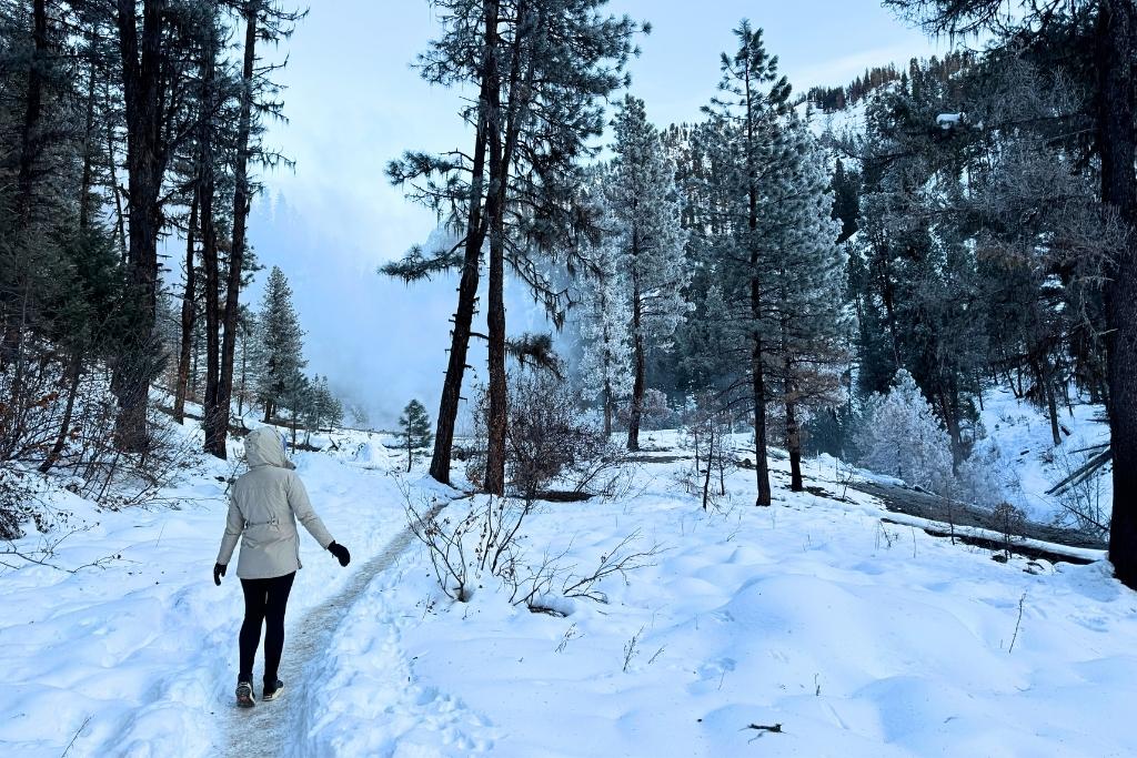 Kate in a white winter coat and black leggings walks along a snowy trail through a frost-covered forest, with steam rising in the distance at Bonneville Hot Springs.