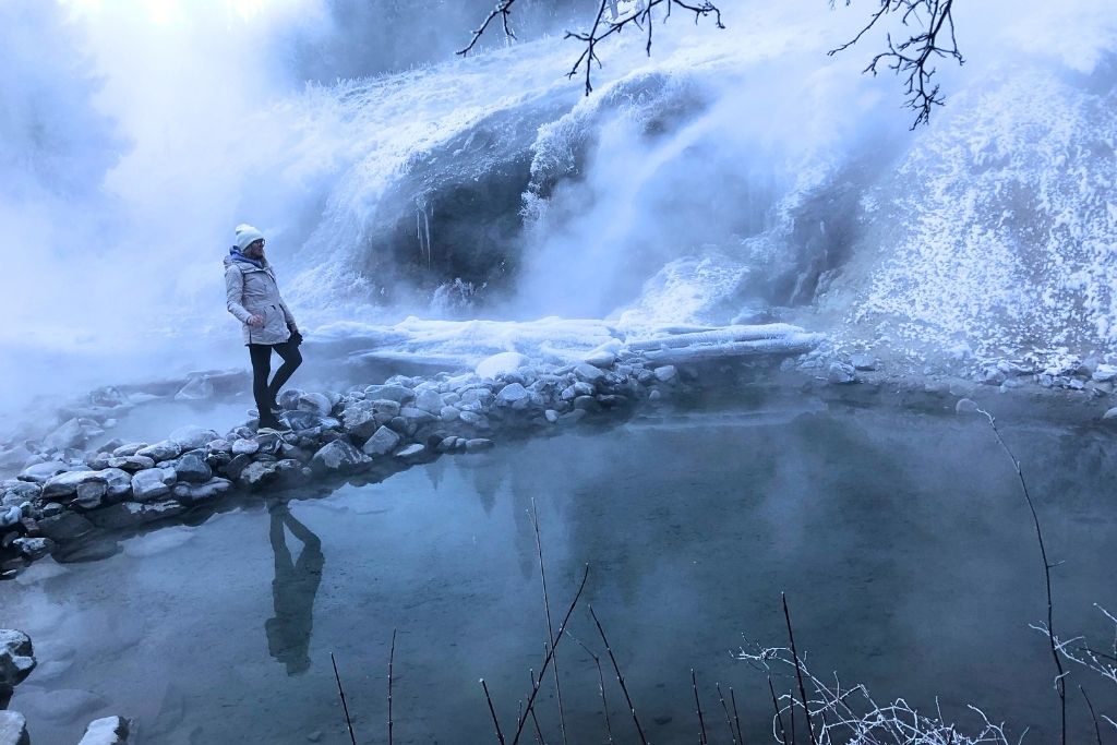 Kate in a white winter coat and beanie walks along a rocky path beside a steaming geothermal pool at Bonneville Hot Springs, with mist rising from the cascading hot water and frost-covered ground surrounding the scene.