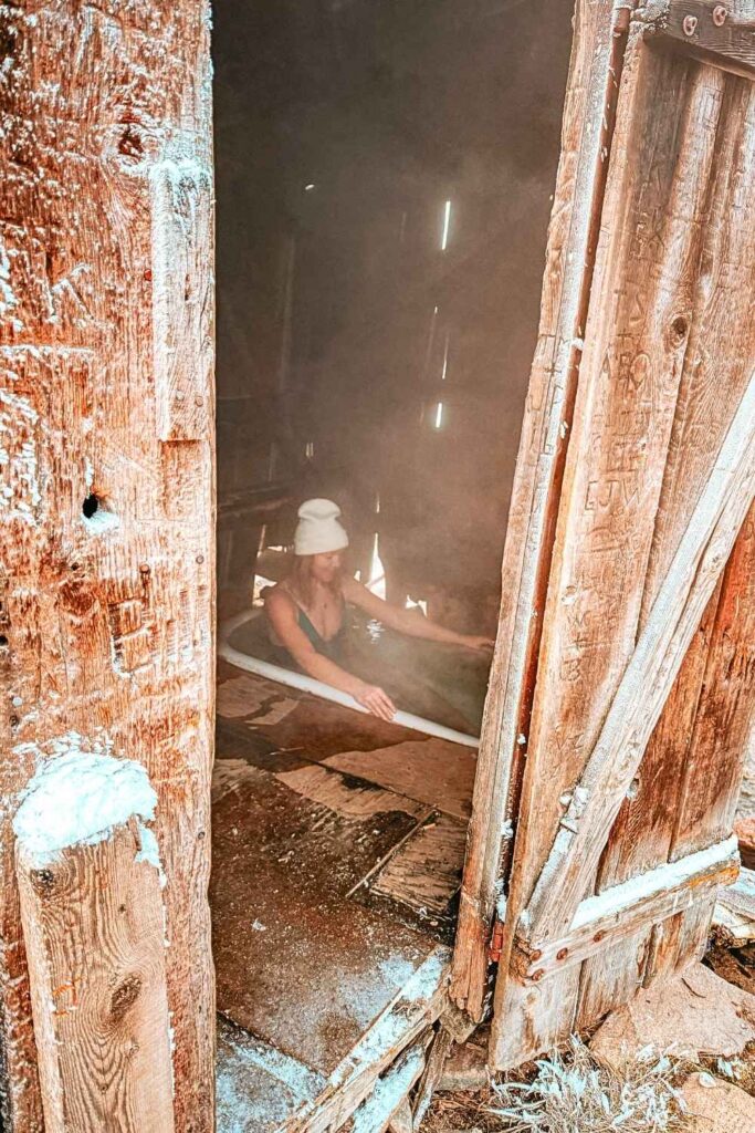 Kate in a green swimsuit and white beanie soaks in a steaming wooden tub inside a rustic, weathered shack at Bonneville Hot Springs, with sunlight filtering through the gaps in the wooden walls.