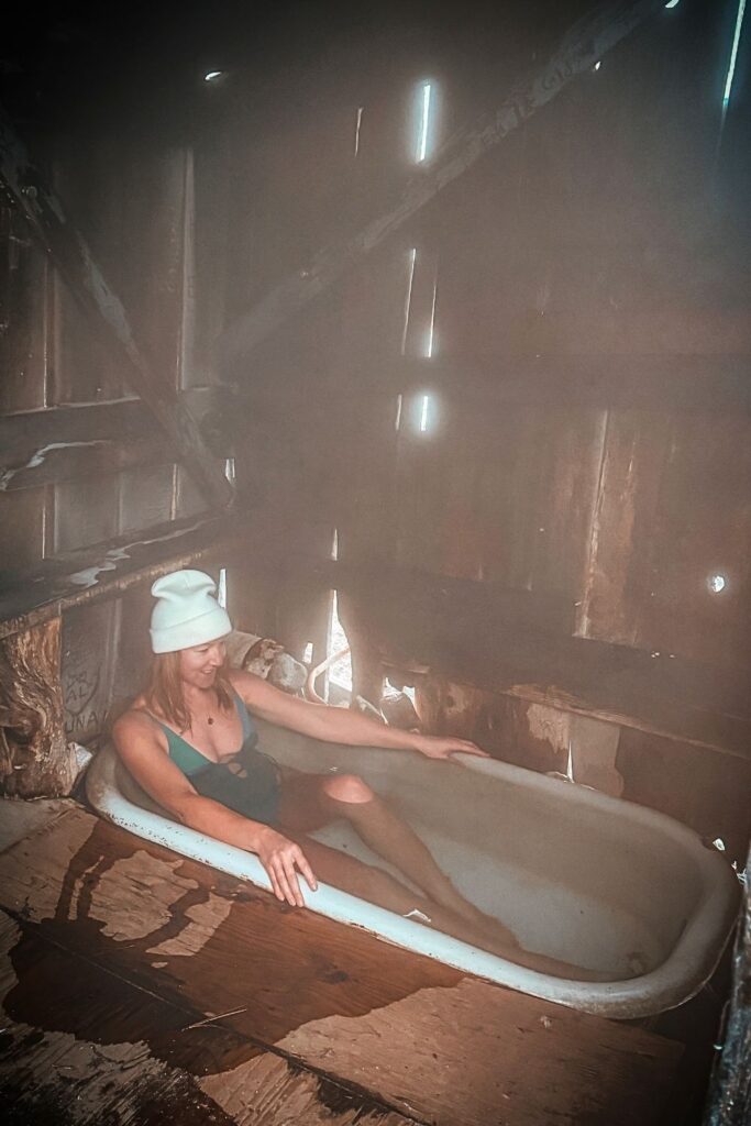 Kate in a green swimsuit and white beanie, relaxes in a steaming old bathtub inside a rustic wooden shack at Bonneville Hot Springs, with mist filling the dimly lit space.