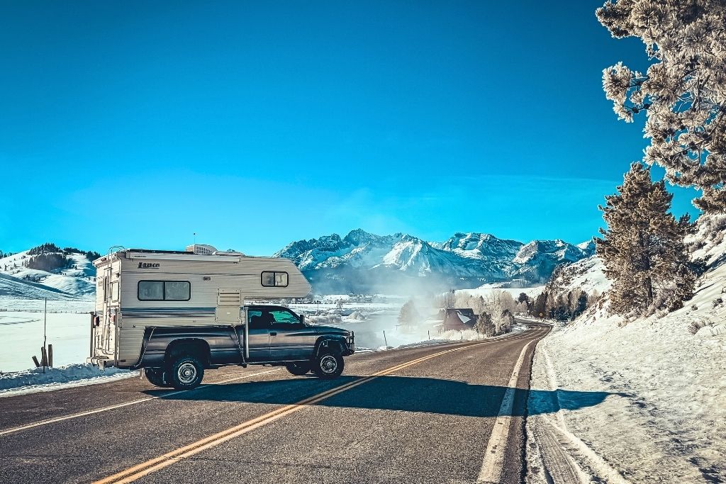 A truck camper is parked on a winding road in Stanley, Idaho, surrounded by snow-covered fields and towering mountain peaks under a bright blue sky, with steam rising from nearby hot springs.