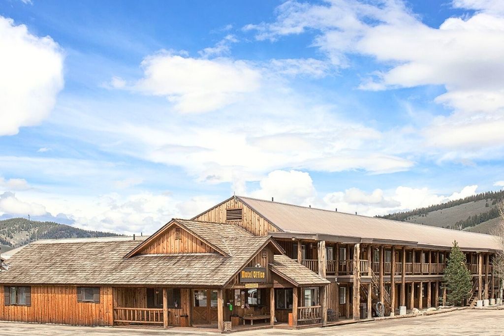 A rustic Mountain Village lodge with a covered porch in Stanley, Idaho sits under a bright blue sky with scattered clouds, surrounded by rolling hills and distant mountains.