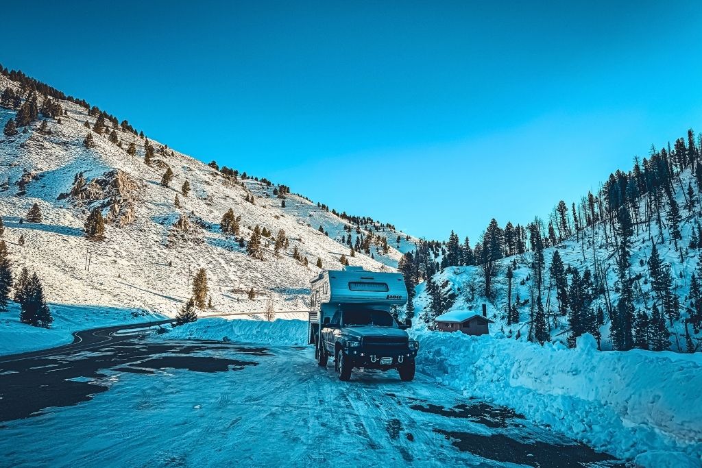 A truck camper is parked on a snow-covered roadside, surrounded by rolling, tree-dotted mountains under a clear blue sky, with a small cabin nestled in the valley below.