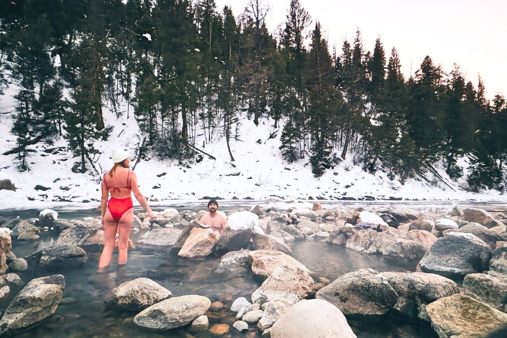 Kate in a red swimsuit and white beanie wades through the steamy Sunbeam Hot Springs, while her husband relaxes in the water, surrounded by snow-covered rocks and a wintery forest backdrop.