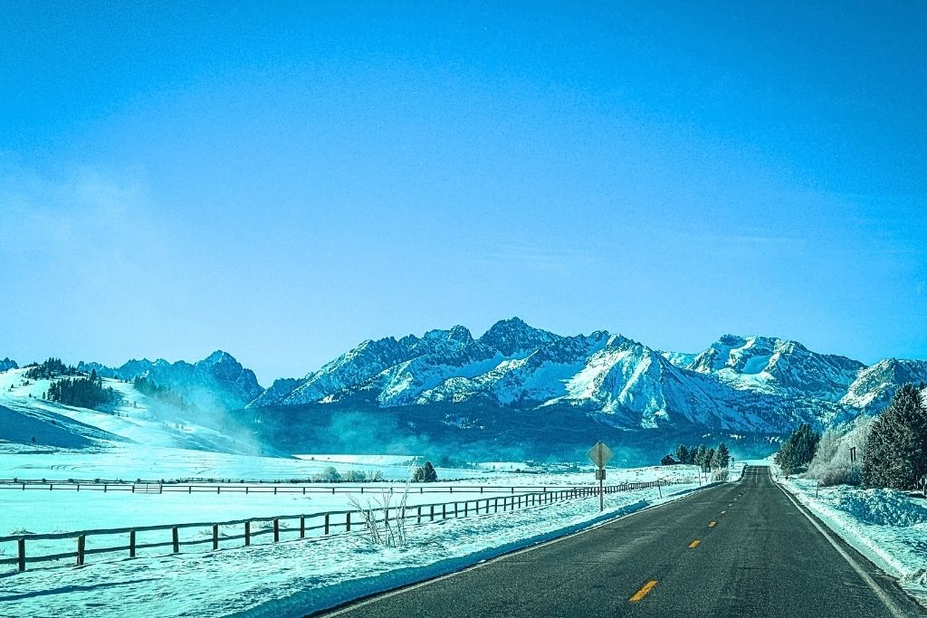 A scenic highway stretches through a snow-covered valley outside Stanley, Idaho, with wooden fences lining the road and the rugged Sawtooth Mountains towering in the background under a clear blue sky.