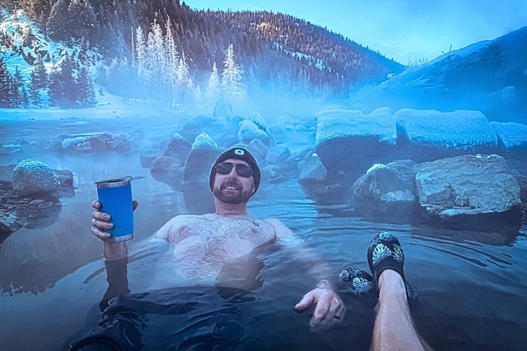 Kate's husband wearing sunglasses and a beanie lounges in a steaming hot spring in Idaho, holding a blue thermos with snow-covered rocks and frost-covered trees in the misty winter landscape around him.