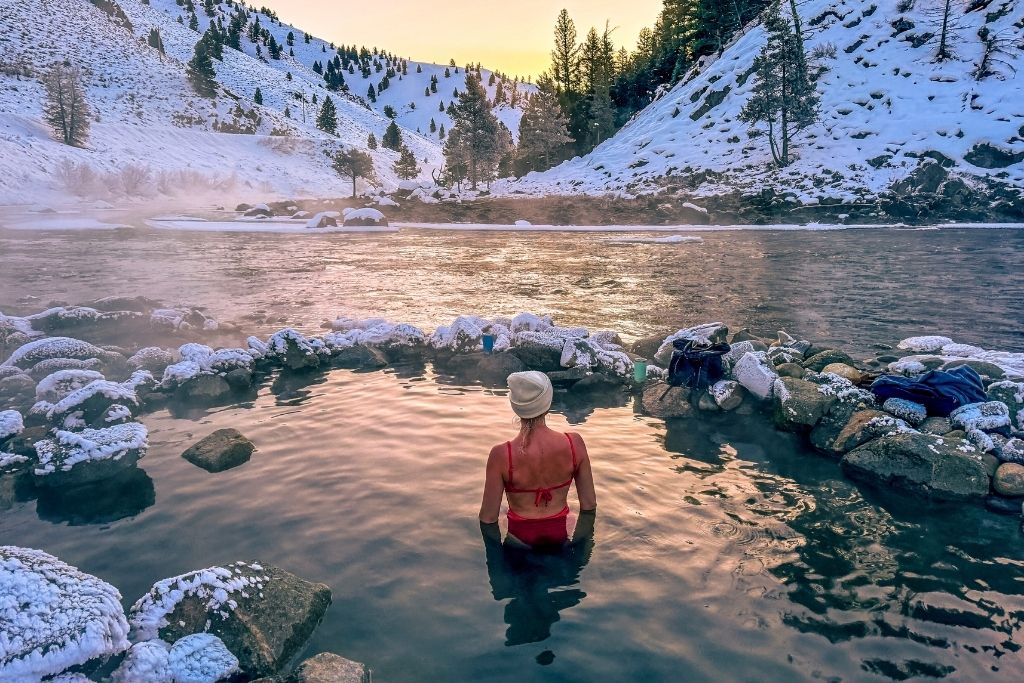 Kate in a red swimsuit and white beanie sits in the steaming Cove Hot Springs, surrounded by snow-covered rocks, with a river and a wintery mountain landscape glowing under the golden light of sunrise.