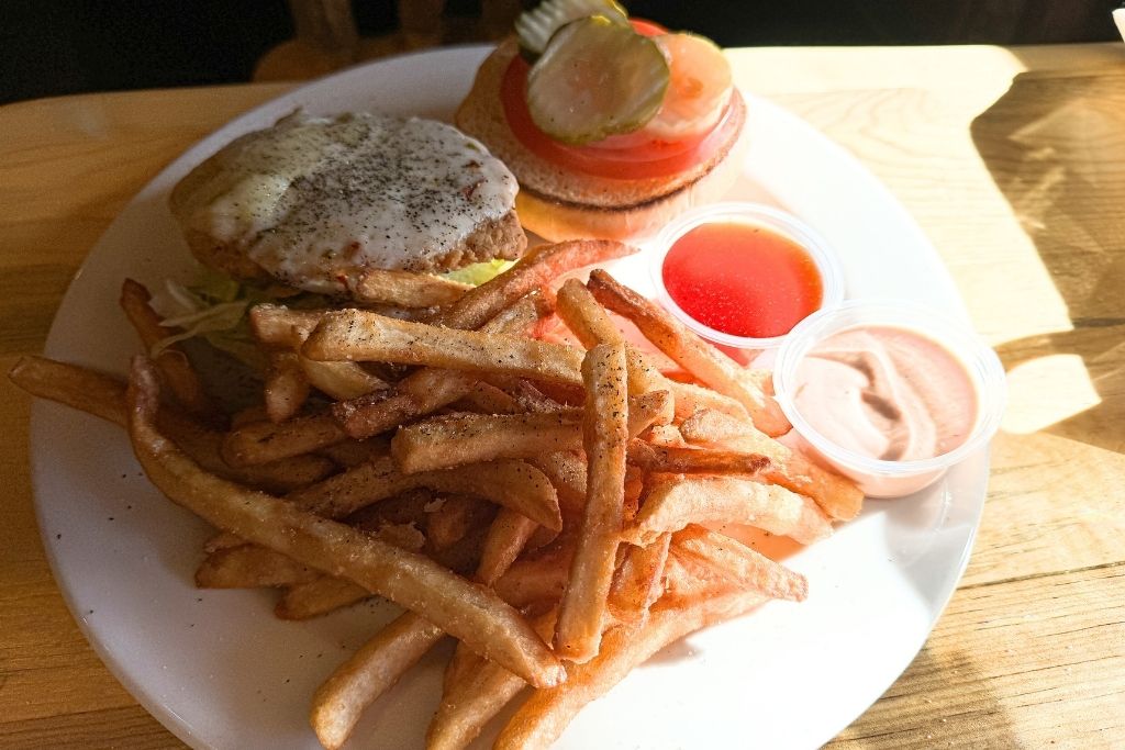 A chicken sandwich with melted cheese, lettuce, tomato, and pickles sits open-faced on a white plate, accompanied by crispy seasoned fries and two dipping sauces on a wooden table.