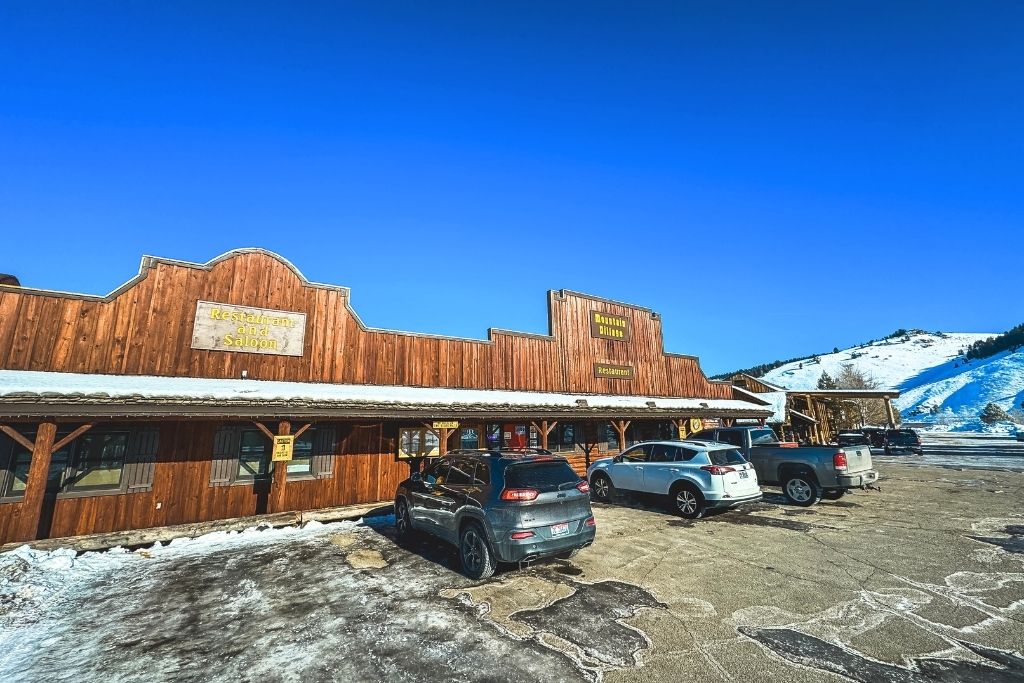 A rustic wooden restaurant and saloon with a Western-style facade sits under a bright blue sky in Stanley, Idaho, with parked cars in front and snow-covered hills in the background.