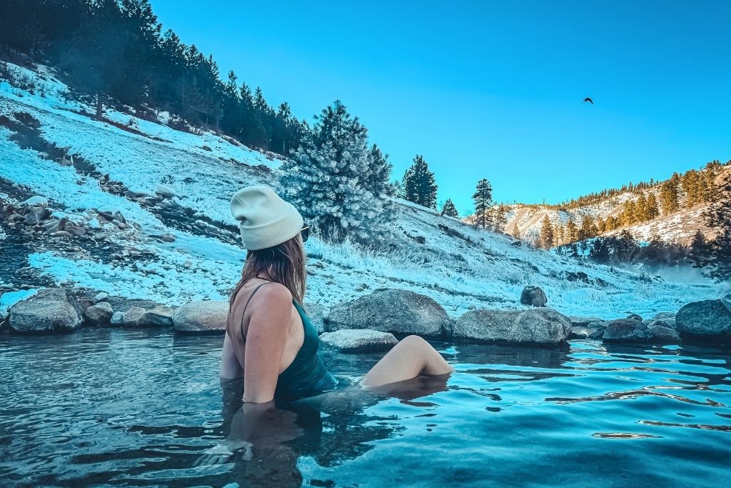 Kate in a green swimsuit and white beanie relaxes in the warm waters of Kirkham Hot Springs, surrounded by snow-covered hills, evergreen trees, and a clear blue sky with a bird soaring overhead.