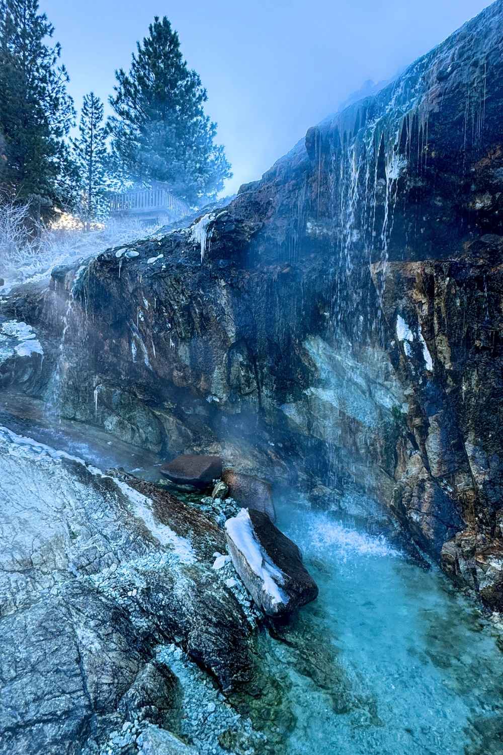 A steaming waterfall cascades over rugged, rocky cliffs into a turquoise pool at Kirkham Hot Springs, with icicles forming on the rocks and mist rising into the crisp winter air.