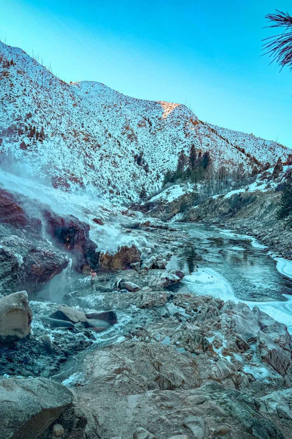 A scenic view of Kirkham Hot Springs with steaming waterfalls cascading over rocky terrain, surrounded by snow-covered mountains and a partially frozen river under a clear blue sky.