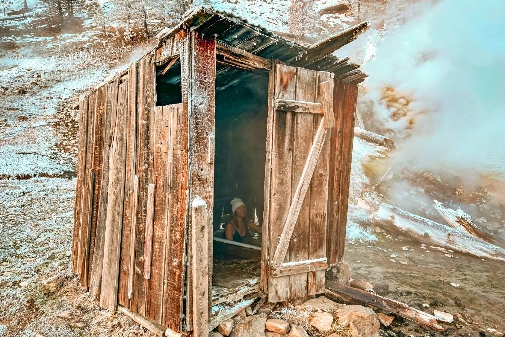 A weathered wooden shack with a partially open door, revealing a rustic interior, stands against a crumbling wall with steam or mist rising around it at Bonneville Hot Springs.