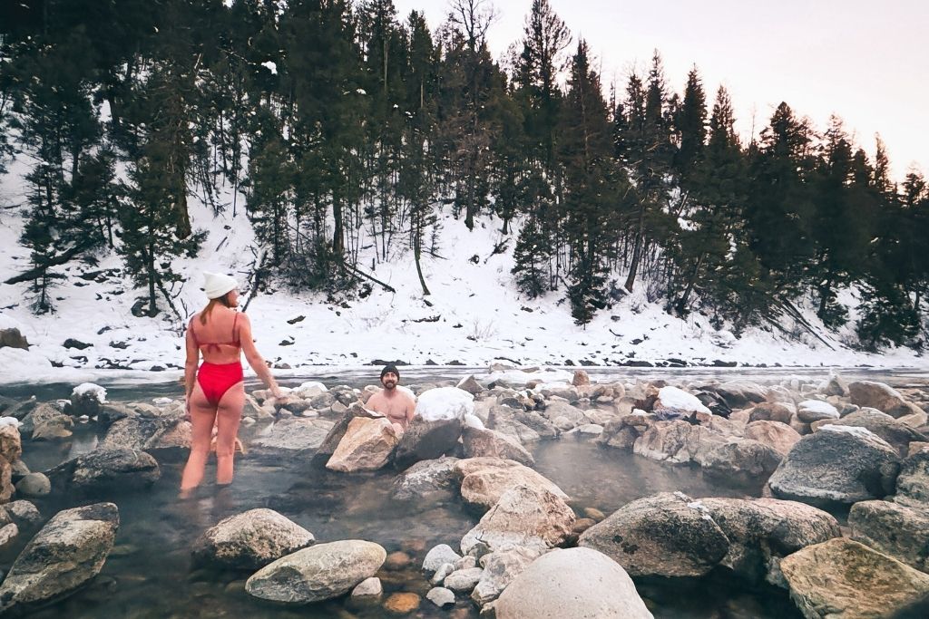 Kate in a red swimsuit and white bucket hat steps through the rocky waters of Sun Beam Hot Springs near Stanley, Idaho, while her husband relaxes in the steaming pool, surrounded by snow-covered rocks and a forested hillside.