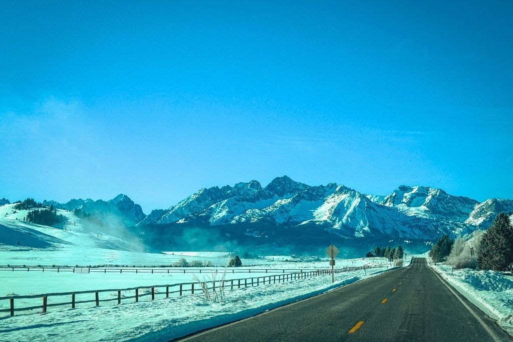 A two-lane road stretches through a snow-covered valley toward the rugged, snow-capped Sawtooth Mountains under a clear blue sky outside Stanley, Idaho.