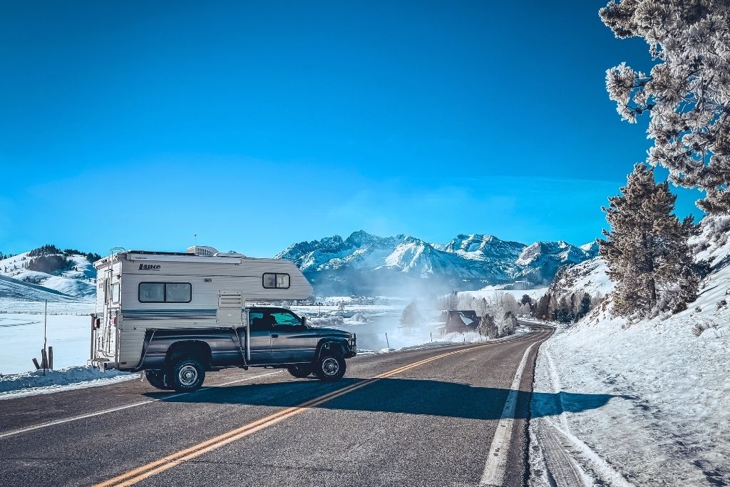 A black pickup truck with a white Lance camper is parked on a snowy mountain road with the rugged, snow-covered Sawtooth Mountains in the background under a clear blue sky.