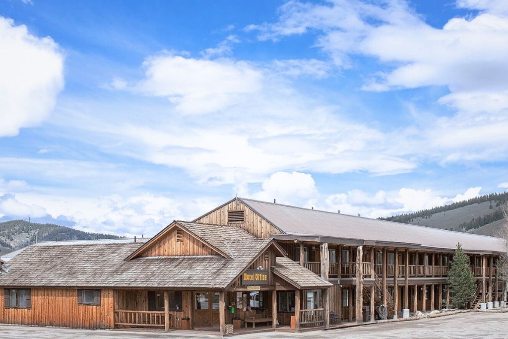 The rustic Mountain Village lodge with a covered porch and log cabin-style architecture stands against a backdrop of rolling hills and a partly cloudy blue sky in Stanley, Idaho.