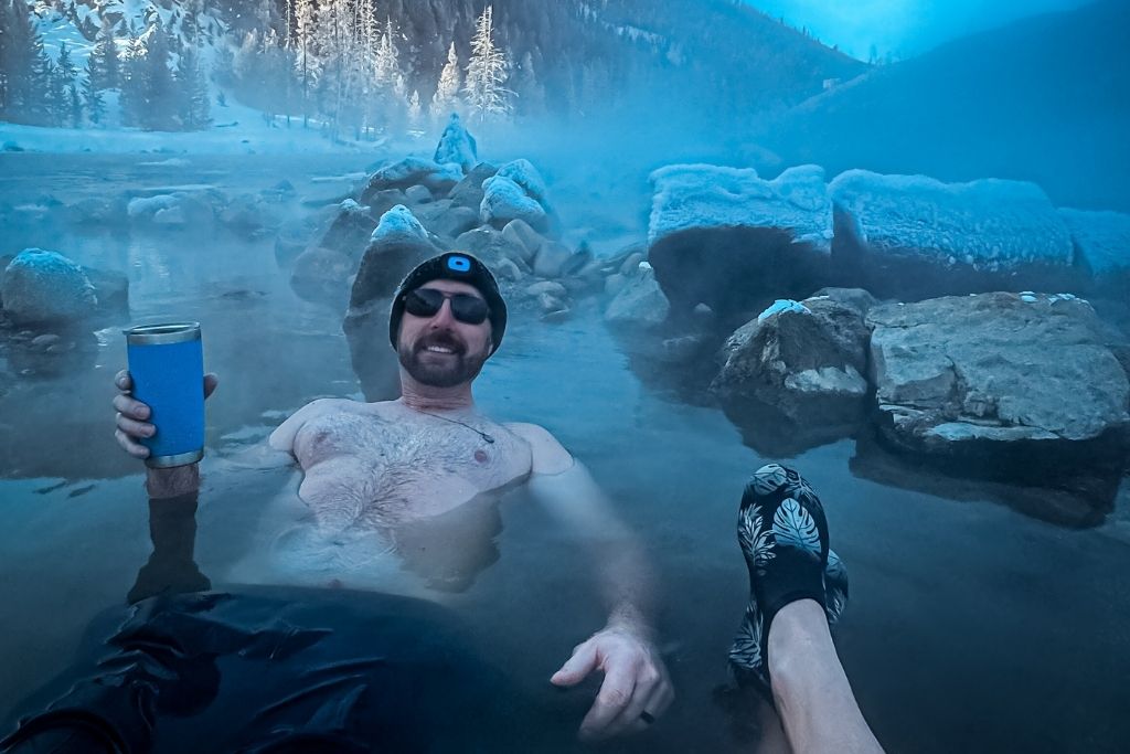 Kate's husband wearing sunglasses and a beanie leans back in a steaming hot spring, holding a blue insulated mug, while Kate's legs rest in the water next to him, surrounded by snow-covered rocks and misty mountain scenery.