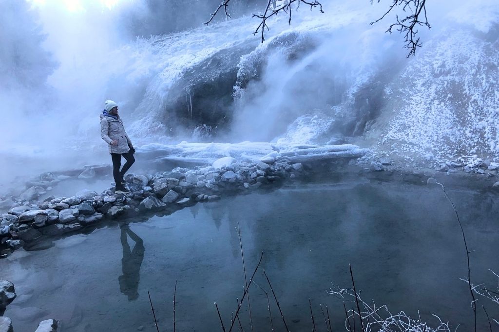 in a winter coat and white beanie walks along a rocky path beside a steaming hot spring at Bonneville Hot Springs, with snow-covered hills and cascading geothermal water in the misty background.