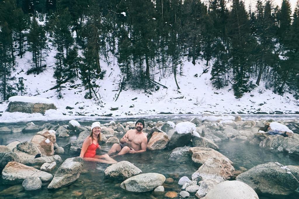 Kate and her husband soaks in the warm waters of Sun Beam Hot Springs, surrounded by large snow-dusted rocks and a backdrop of a snow-covered hillside with evergreen trees.